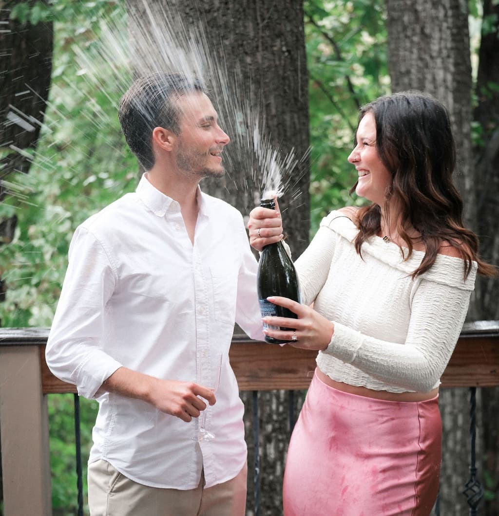 Caroline and Chandler laughing as a champagne bottle sprays between them on a wooded deck