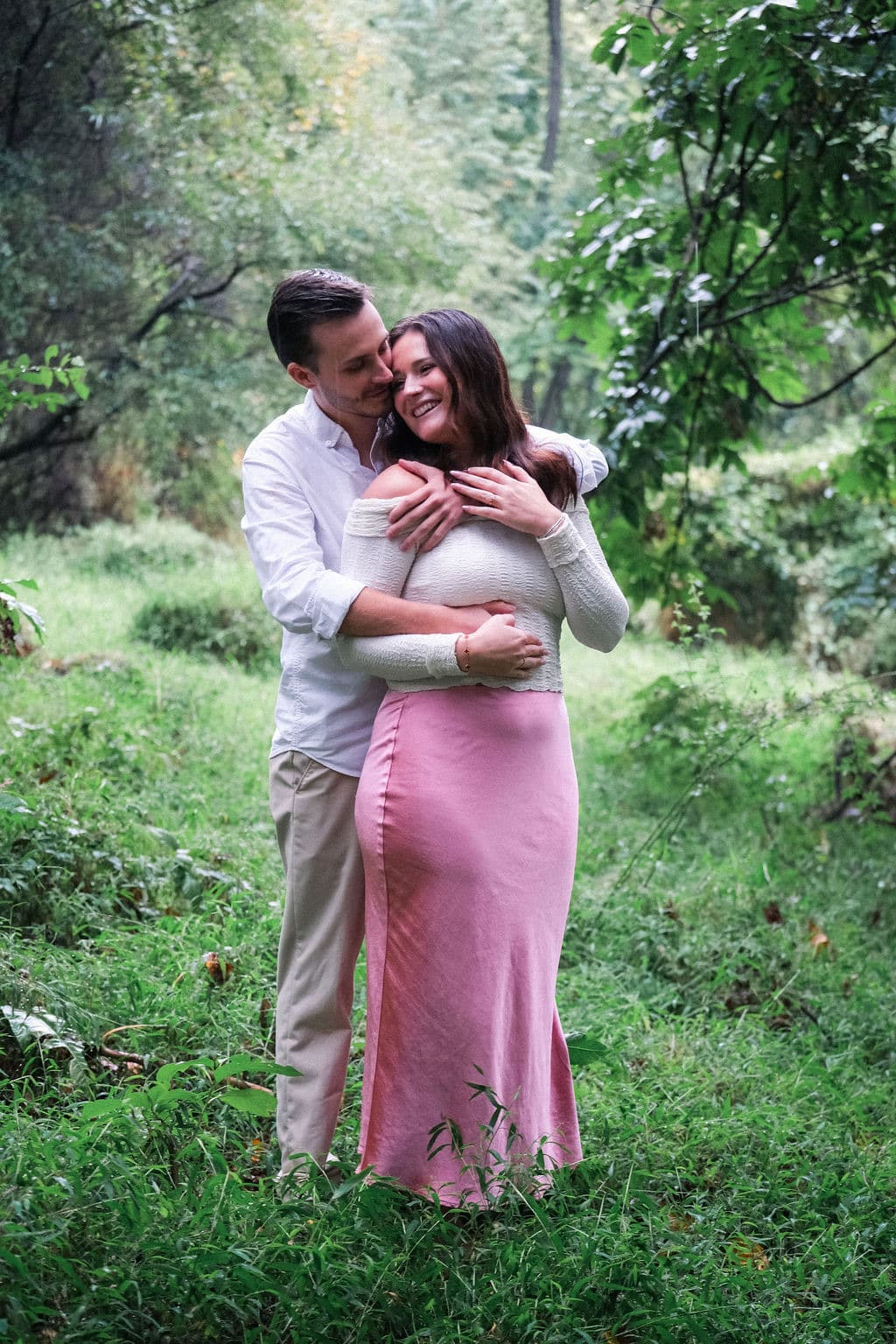 Chandler hugging Caroline from behind in a green clearing